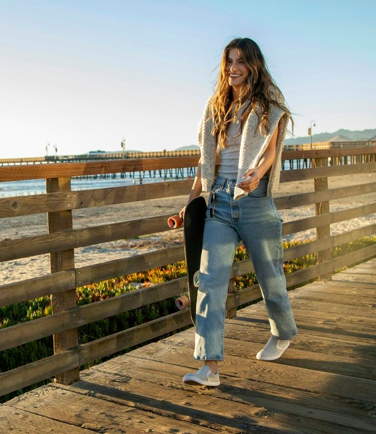 A woman wearing Dillon Canvas Slip-ons walking along the board walk with her skateboard and sweater