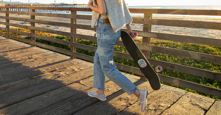 A woman walking down a boardwalk with a skateboard in hand while wearing the Dillon Canvas Slip-On in Lunar Rock